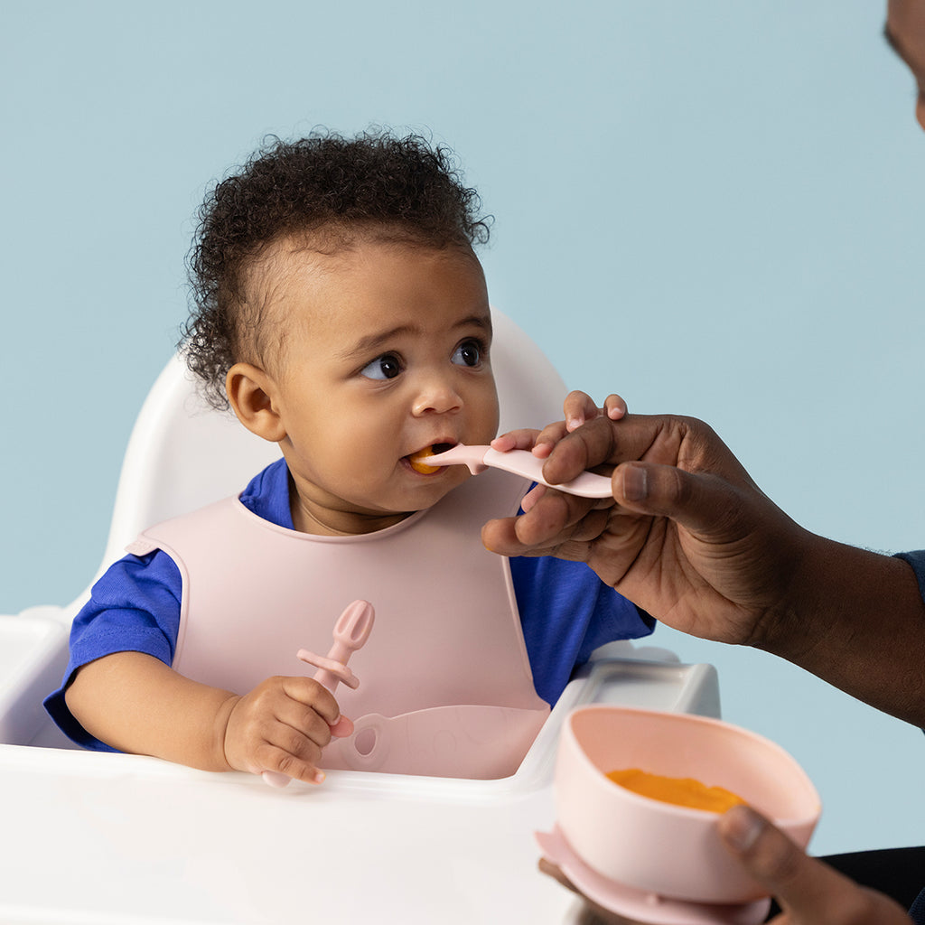 Baby being fed with a spoon by an adult against a light blue background