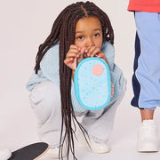 Three children in casual clothing pose with colorful lunch bags and containers in a bright studio setting. One child crouches holding a light blue lunch bag, while two standing children hold blue and green lunch containers.
