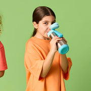 Three children standing against a solid green studio background, each holding and drinking from a reusable water bottle with a blue cap and teal bottle body.