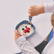 Child eating yogurt with berries from a blue bowl on a white table.