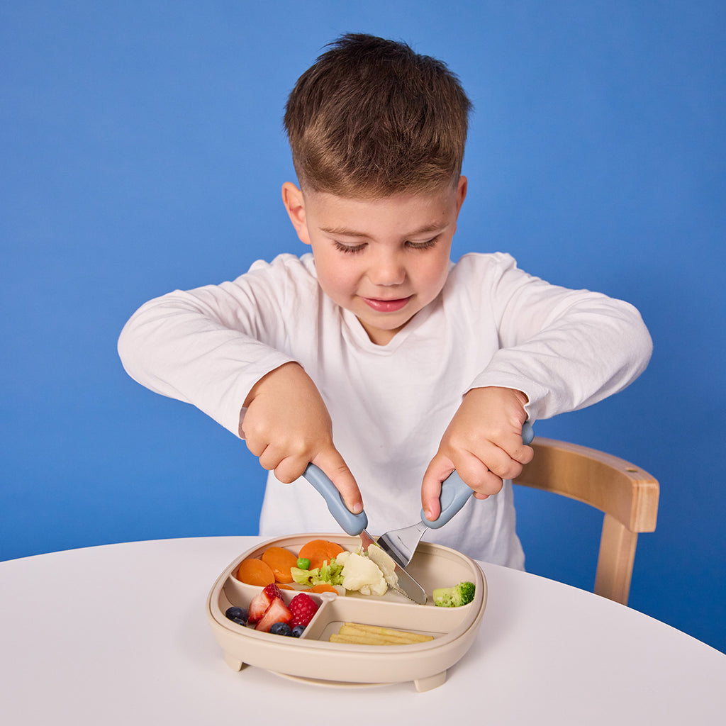 Child using a knife to cut food on a divided plate against a blue background