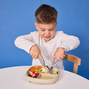 Child using a knife to cut food on a divided plate against a blue background