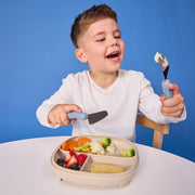 Child with a plate of vegetables and fruits, holding a fork and spoon against a blue background