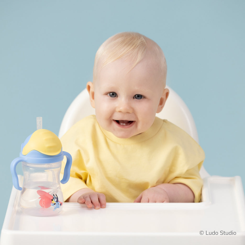 Baby in a high chair with a yellow top and a Bluey sippy cup on a light blue background