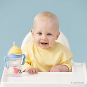Baby in a high chair with a yellow top and a Bluey sippy cup on a light blue background