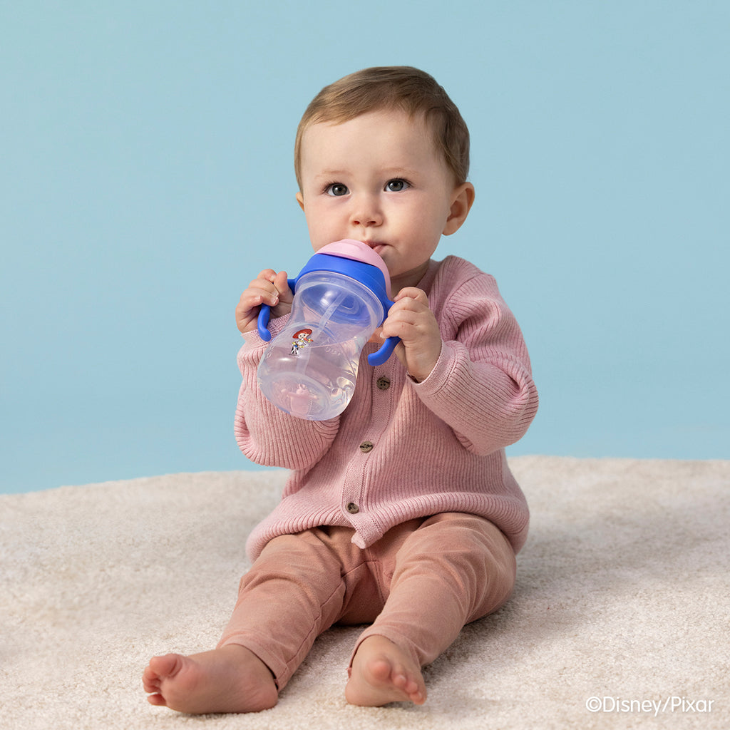 Baby in a pink outfit holding a blue sippy cup against a light blue background