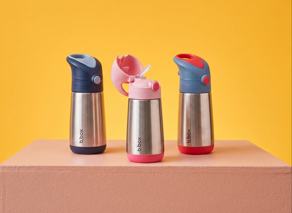 Three aBox insulated water bottles with colorful lids are displayed on a pink pedestal against a solid yellow background. The bottles feature stainless steel bodies and lids in blue, pink, and red colorways.